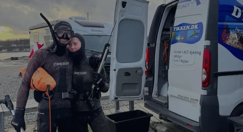 Deux plongeurs en combinaison, souriants, devant un van avec du matériel