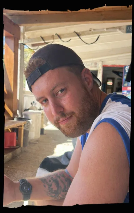 Portrait d'un homme barbu souriant, casquette, devant un abri