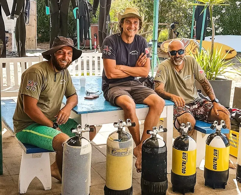 Trois plongeurs souriants assis avec des bouteilles devant un bâtiment