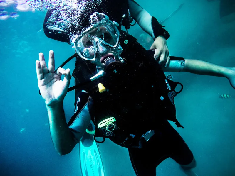 Plongeur souriant avec signe OK, équipé, dans une eau turquoise