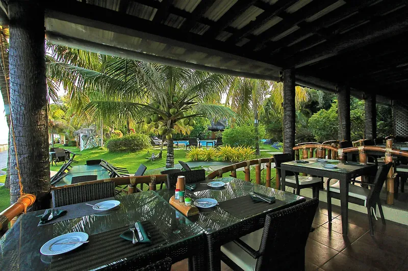 Terrasse ombragée avec tables dressées, vue sur jardin tropical et mer