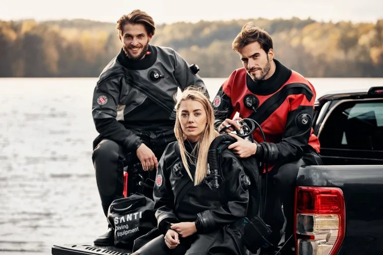 Trois plongeurs souriants en combinaison près d'un véhicule et de l'eau