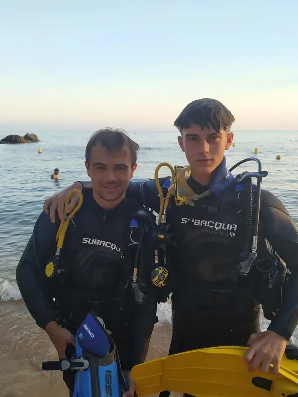 Deux plongeurs souriants en combinaison, équipés, sur une plage ensoleillée