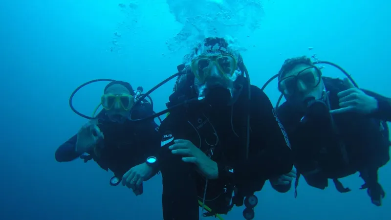 Trois plongeurs en combinaison, masques et détendeurs, sourient sous l'eau
