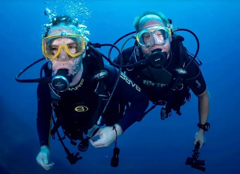 Deux plongeurs souriants en combinaison, équipés, nagent dans l'eau bleue
