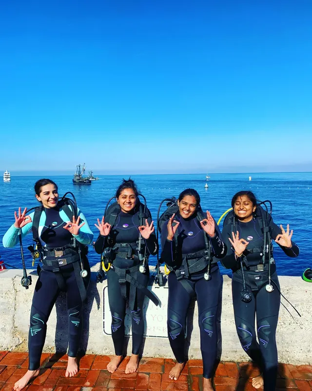 Quatre plongeuses souriantes en combinaison, équipées, sur un quai ensoleillé