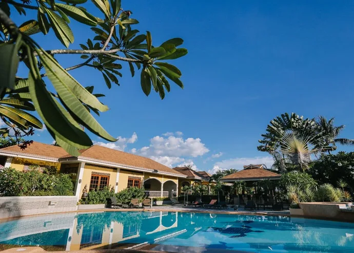 Piscine devant bâtiments colorés, végétation luxuriante et ciel bleu clair