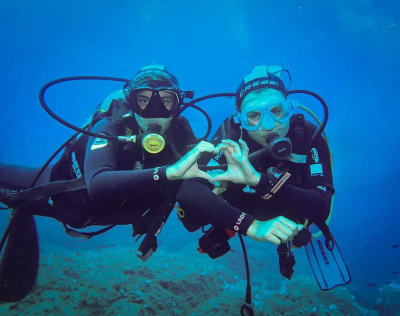 Deux plongeurs souriants, équipés, font un signe de la main sous l'eau