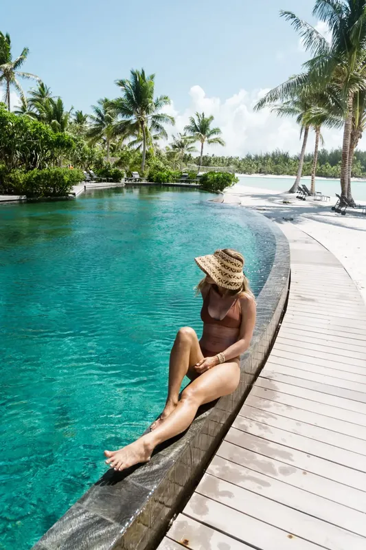 Femme assise au bord d'une piscine turquoise avec un chapeau