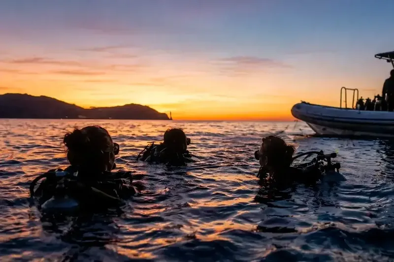 Trois plongeurs équipés nagent vers un bateau au coucher du soleil