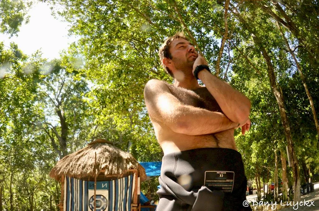 Homme torse nu, regard pensif, devant une structure en bois et végétation