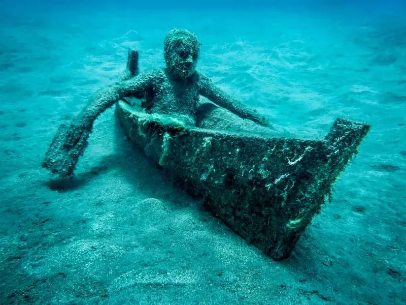 Sculpture sous-marine d'une personne dans un petit bateau, sable bleu