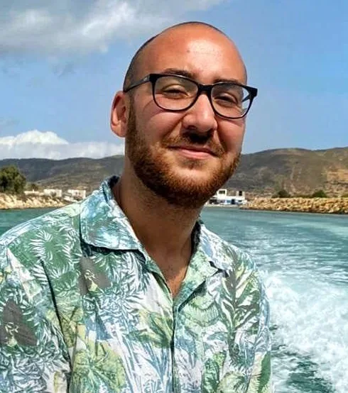 Homme souriant avec lunettes, en chemise, devant la mer et montagnes