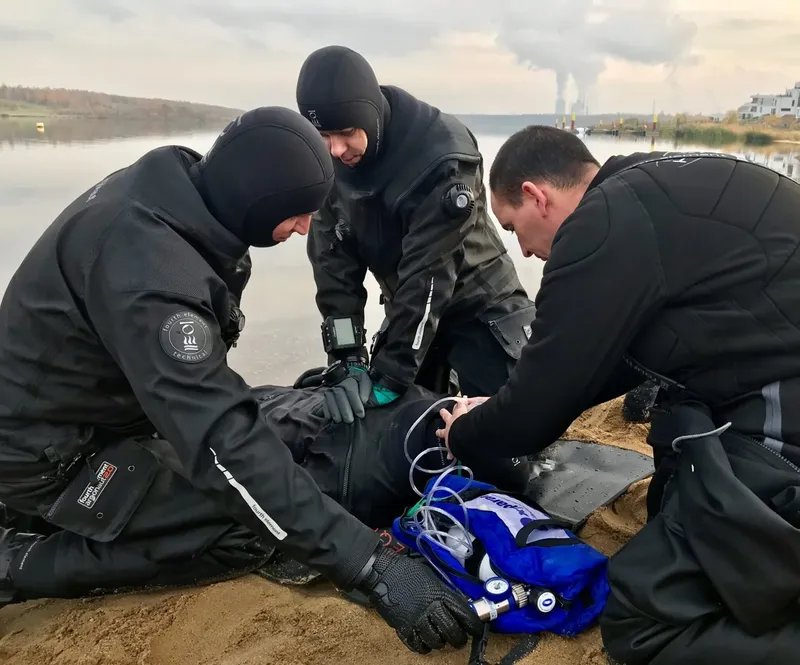 Trois plongeurs en combinaison sèche examinent un appareil sur la plage