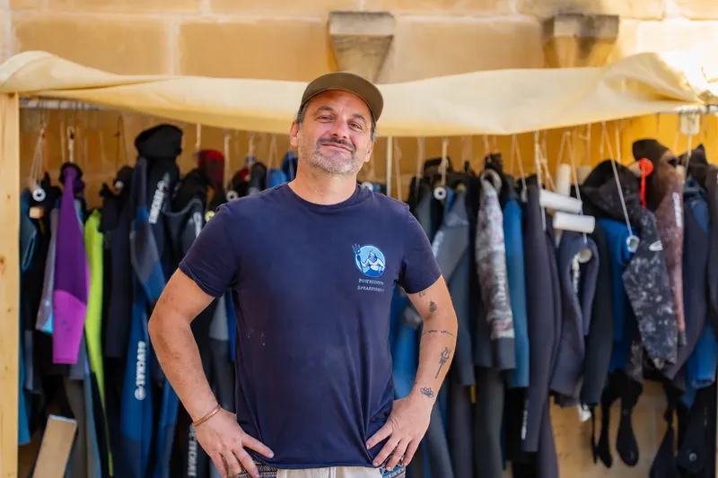 Un homme souriant se tient devant des combinaisons de plongée suspendues