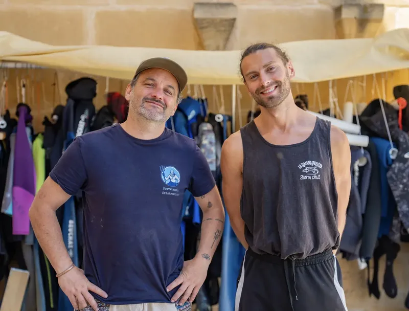 Deux hommes souriants posent devant des combinaisons de plongée et du matériel