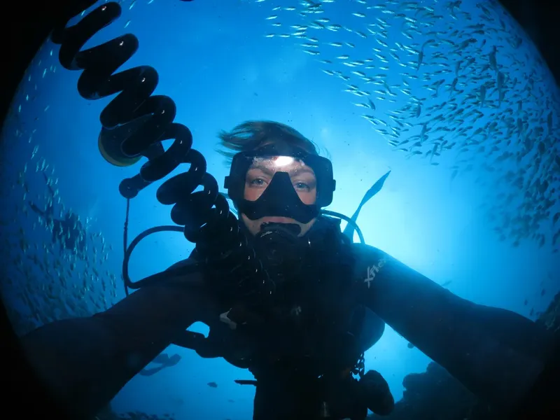 Plongeur souriant face à la caméra, entouré d'un banc de poissons