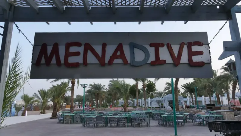 Enseigne lumineuse MENADIVE au-dessus d'une terrasse avec tables et palmiers