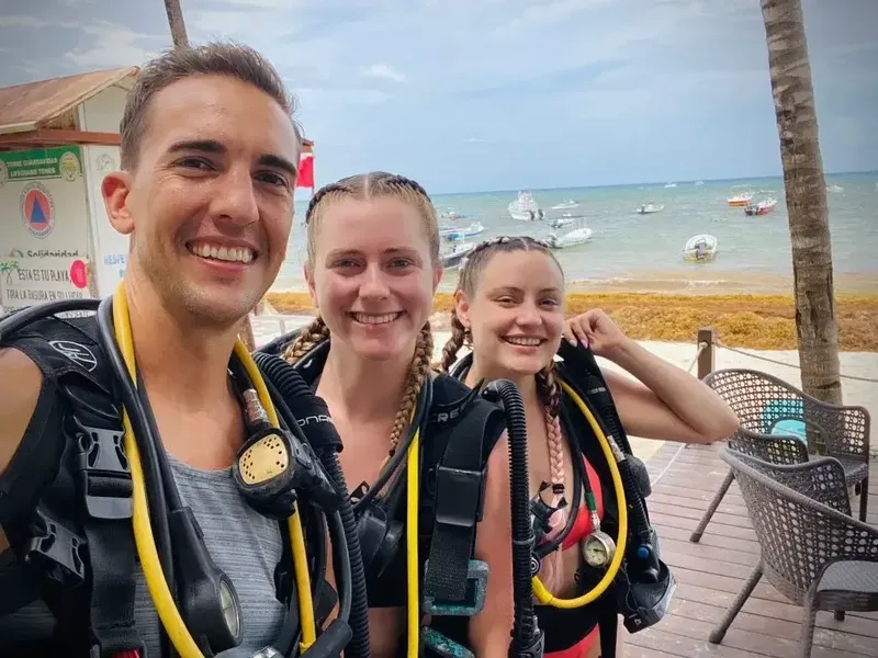 Trois plongeurs souriants équipés devant une plage ensoleillée