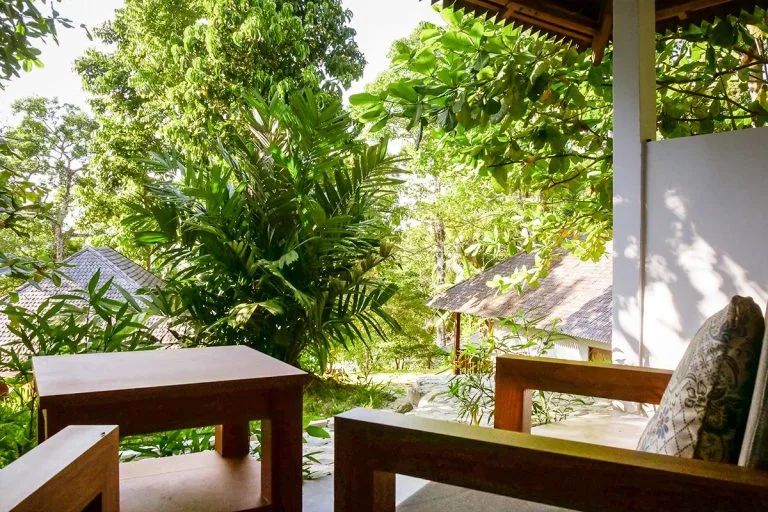 Terrasse en bois avec mobilier, végétation luxuriante et bâtiments en arrière-plan