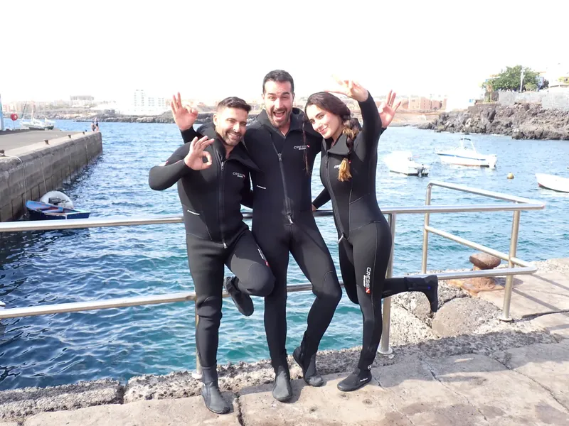 Trois plongeurs souriants en combinaison, bras levés, sur un quai