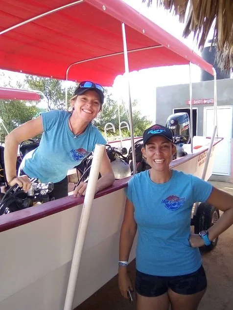 Deux femmes souriantes posent près d'un bateau de plongée et d'un bâtiment