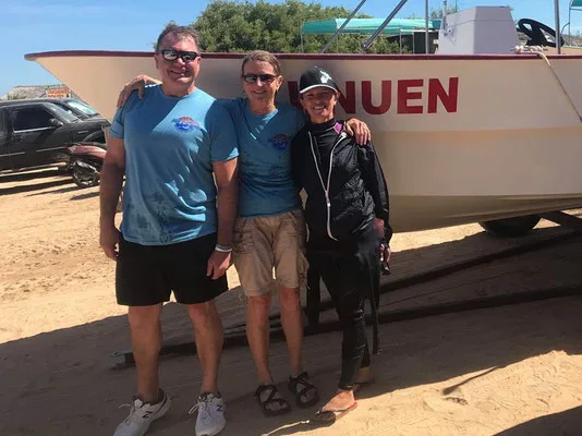 Trois personnes souriantes posent devant un bateau blanc sur une plage
