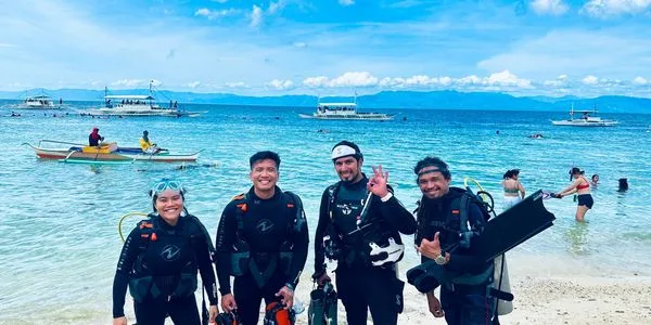 Quatre plongeurs souriants sur une plage avec des bateaux en arrière-plan