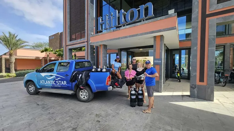 Groupe de plongeurs avec équipement devant un pick-up bleu et un hôtel