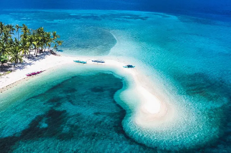 Plage de sable blanc, eau turquoise, quelques bateaux et palmiers visibles