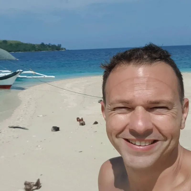 Homme souriant sur une plage paradisiaque avec des bateaux au loin
