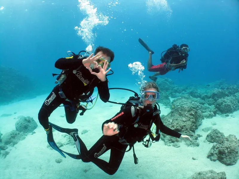 Trois plongeurs souriants font des signes sous l'eau au milieu des coraux