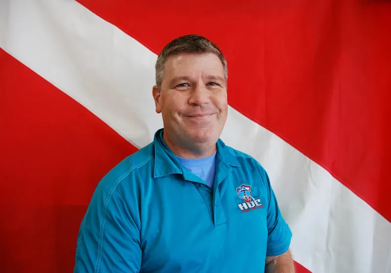 Homme souriant portant un polo bleu devant un drapeau plongee