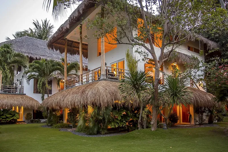 Bâtiment blanc avec toits de chaume, végétation tropicale et pelouse verte