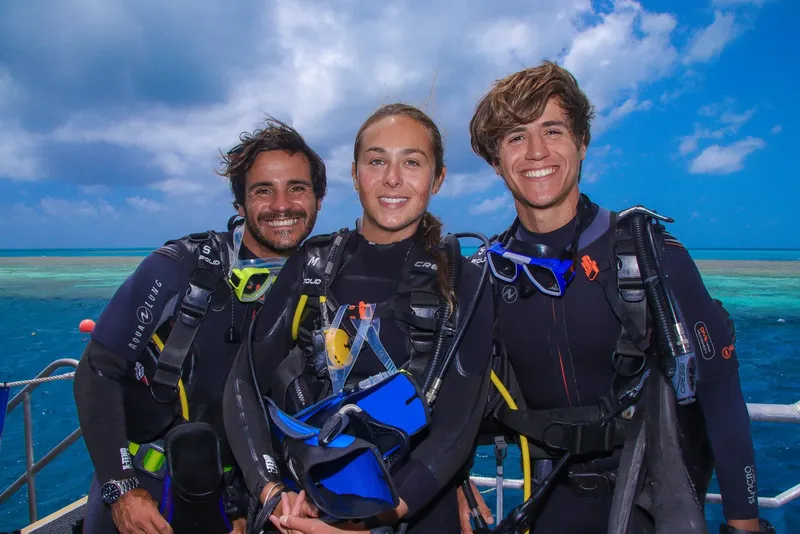 Trois plongeurs souriants en combinaison, équipés, sur un bateau