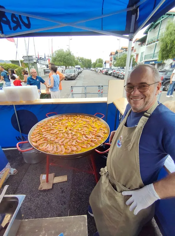 Un homme souriant présente une grande paella sur un stand extérieur