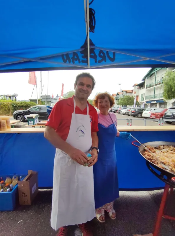 Deux personnes souriantes devant un stand avec une paella et des voitures