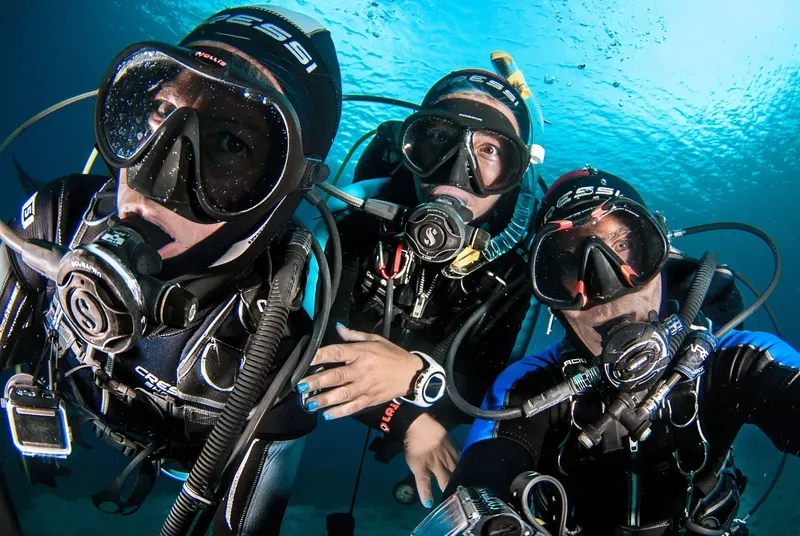 Trois plongeurs souriants, équipés, regardent vers la caméra sous l'eau