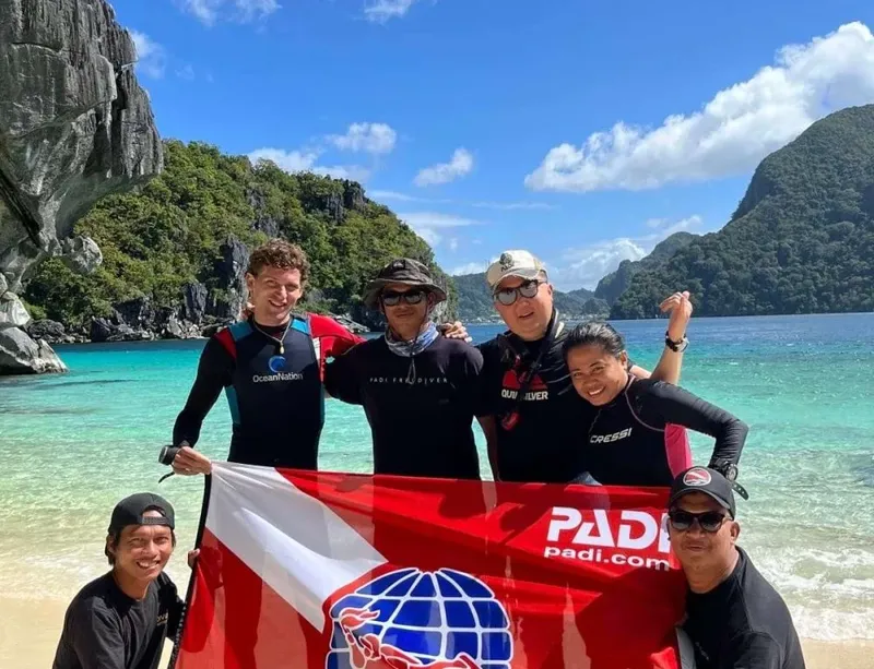 Groupe de plongeurs souriants tenant un drapeau PADI sur une plage