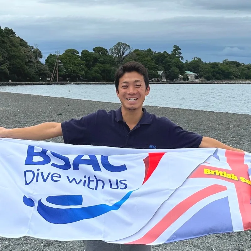 Un homme souriant tient un drapeau BSAC avec un motif de plongée