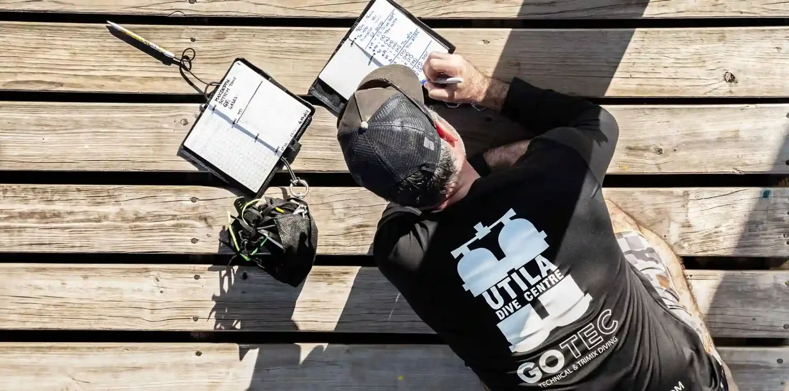 Homme en t-shirt de plongée écrit des notes sur un ponton en bois