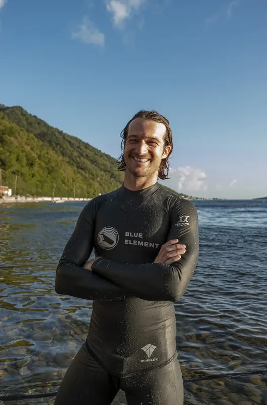 Un homme souriant en combinaison de plongée, bras croisés, au bord de l'eau