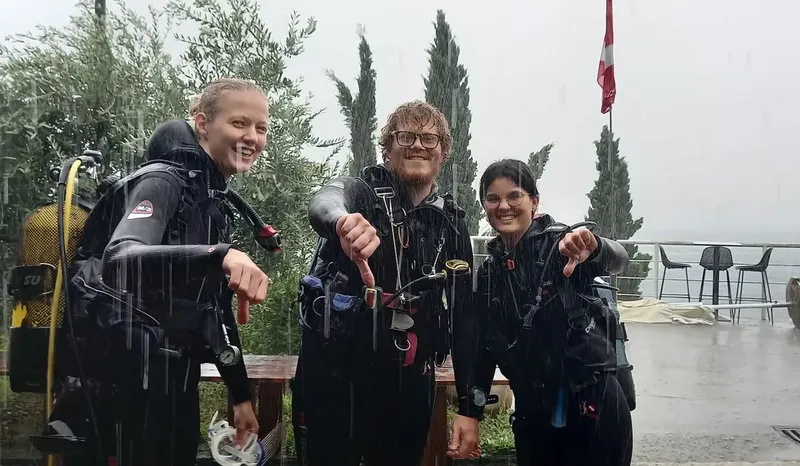 Trois plongeurs souriants en combinaison, équipés, posent sous la pluie