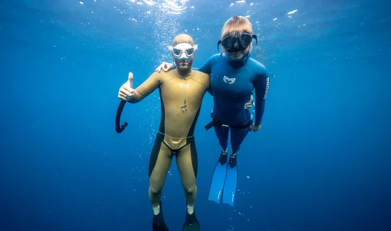 Deux plongeurs souriants en combinaison, pouces levés, dans l'eau bleue
