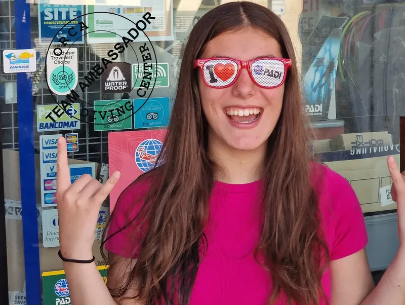 Jeune femme souriante avec des lunettes rouges devant un centre de plongée