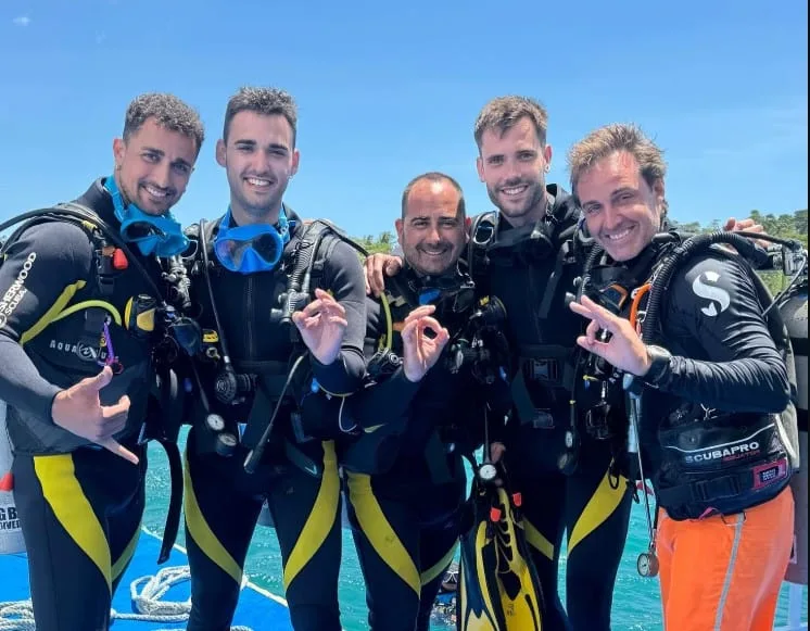 Cinq plongeurs souriants en combinaison, équipés, sur un bateau en mer
