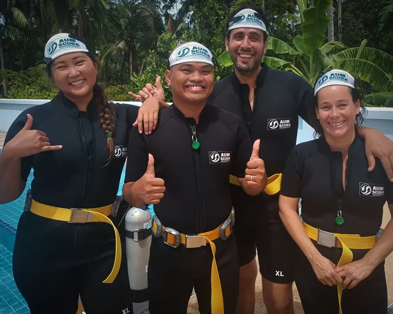 Quatre personnes souriantes en combinaison de plongée avec logo AUM SCUBA