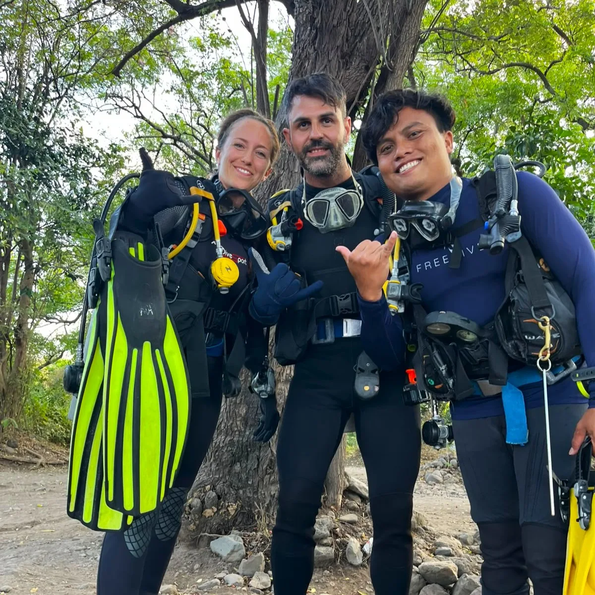 Trois plongeurs souriants en combinaison, équipés, posent sur un sentier rocheux