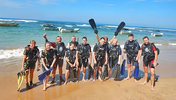 Groupe de plongeurs souriants sur une plage, équipés et prêts à plonger