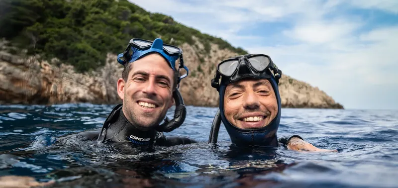 Deux plongeurs souriants émergent de l'eau, paysage côtier en arrière-plan